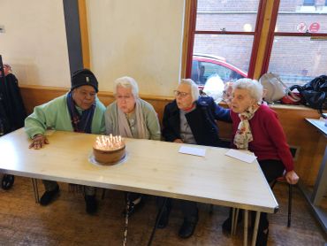 Four elderly people gathered around a birthday cake with lit candles.