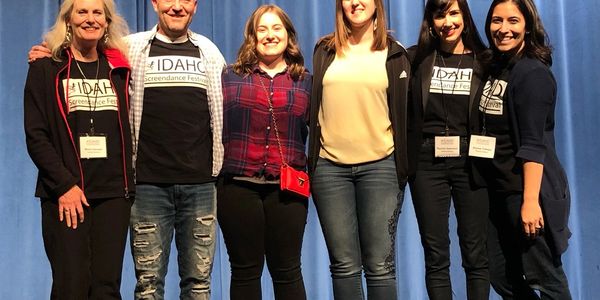 Group photo of six people smiling on stage with blue curtain.