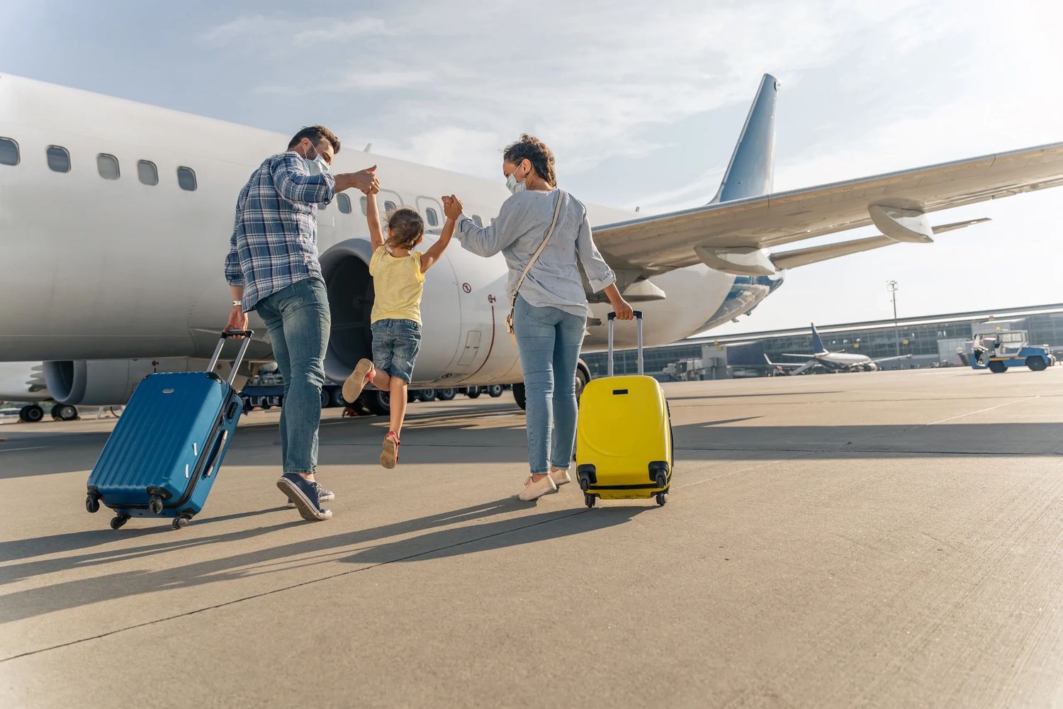 Family with luggage walking to board a plane, holding hands and wearing masks.