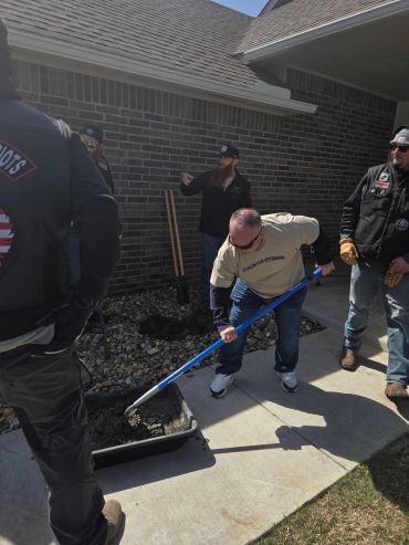 A man shovels concrete while others watch outside a brick house.