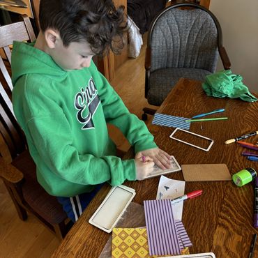 A boy in a green hoodie writing on a small paper at a wooden table with crafting supplies.