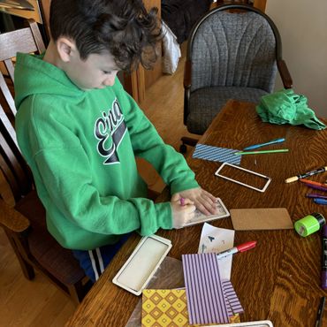 A boy in a green hoodie writing on a small paper at a wooden table with crafting supplies.