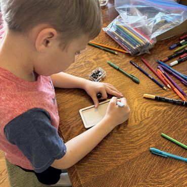 A boy draws on a small whiteboard surrounded by markers and colored pencils.