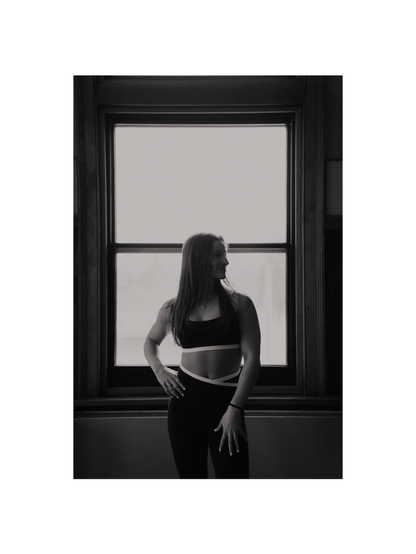 Black and white photo of a woman in athletic wear standing by a window.