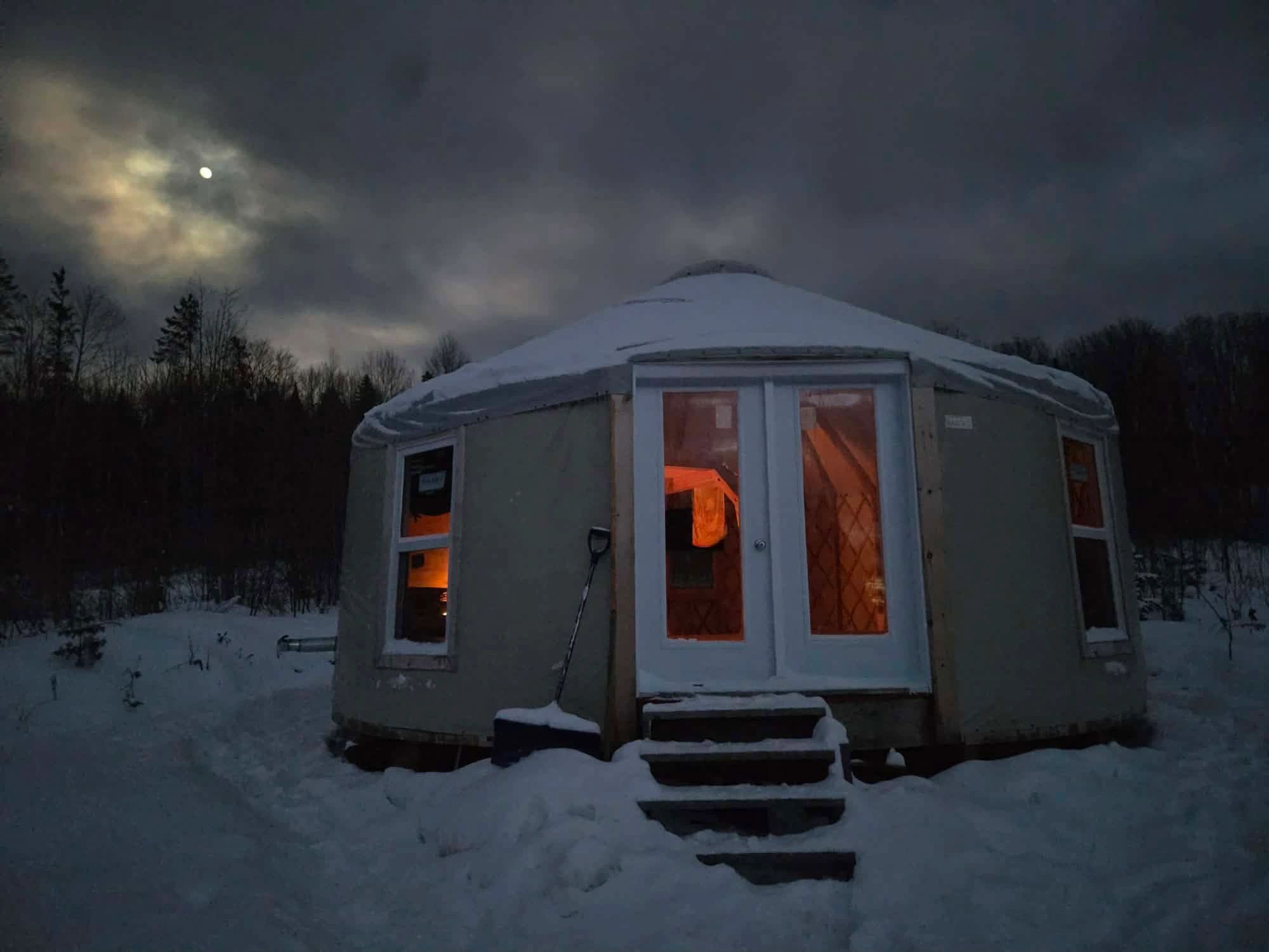A yurt in snowy woods at night glowing warmly inside.