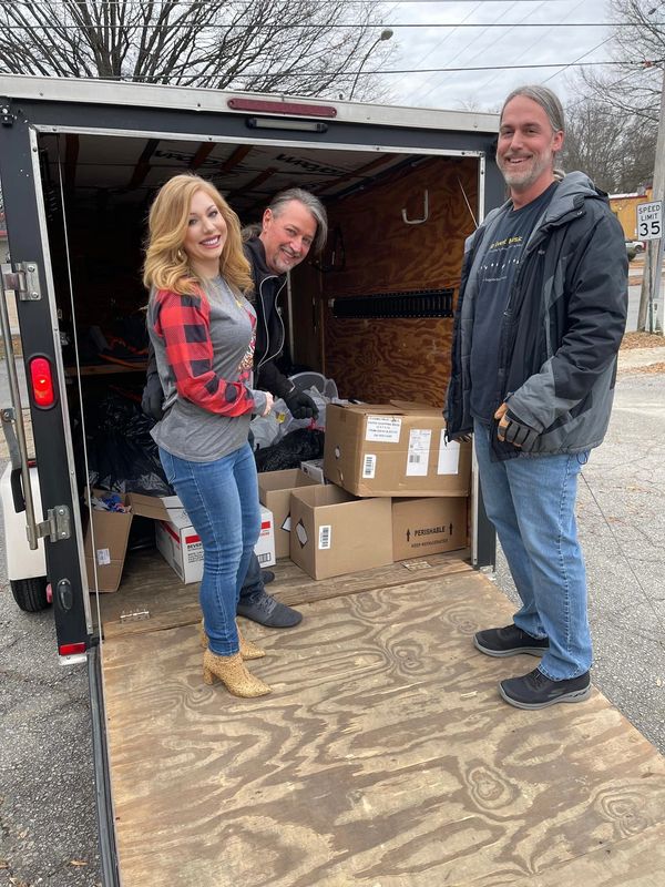 Three people smiling while unloading boxes from a trailer on a cloudy day.