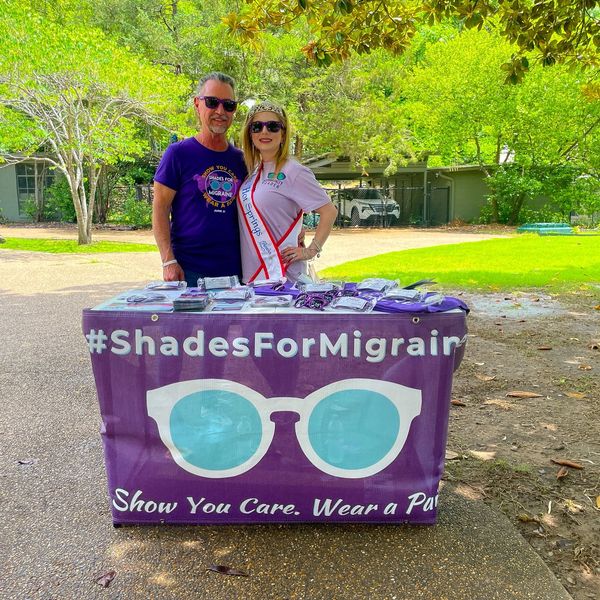 Two people standing behind a table with a #ShadesForMigraine banner outdoors.