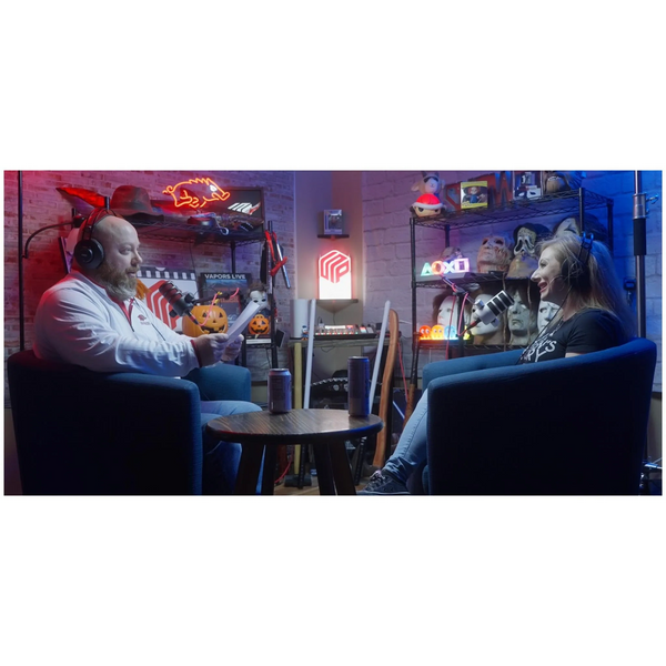 Two people recording a podcast in a Halloween-themed studio with neon lights and masks.