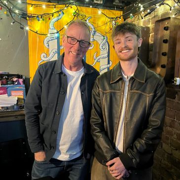 Two men smiling indoors with festive string lights behind them.