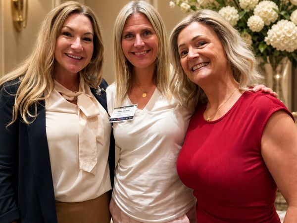 Three smiling women posing indoors with floral decorations in the background.