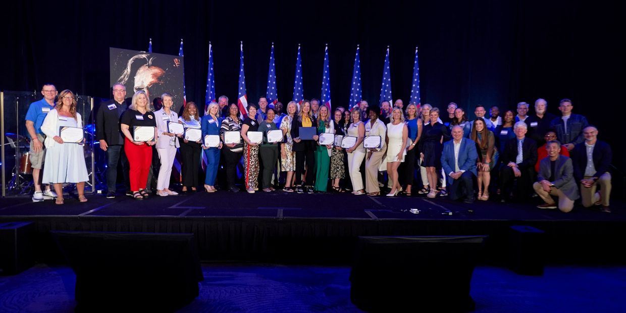 Group photo of diverse individuals holding certificates on stage with American flags behind them.