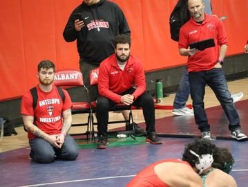 Coaches and a team member watching a wrestling match in a gymnasium.