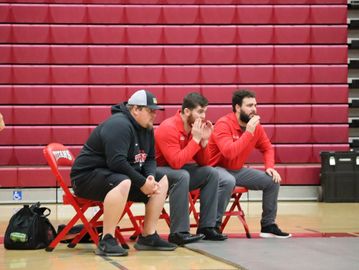 Three men intently watching a wrestling match from red chairs in a gymnasium.
