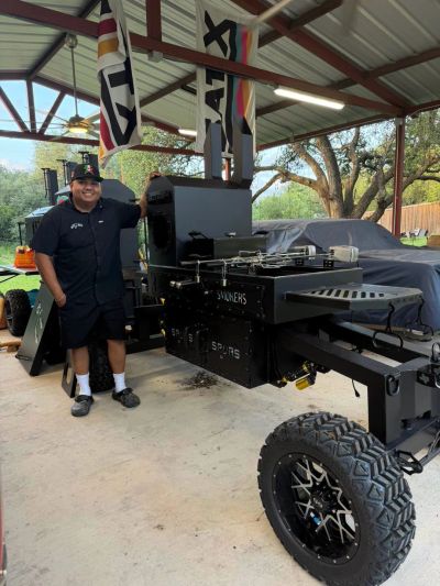 Man standing next to a large smoker grill under a covered patio.