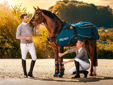Two riders attending a horse outfitted with protective gear in a sunlit outdoor setting.