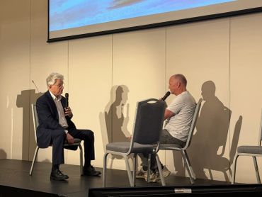 Two men sit on stage, each holding a microphone, engaged in conversation.