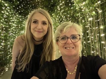 Two women smiling in front of a backdrop of string lights and greenery.
