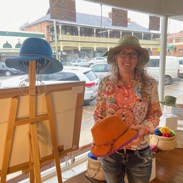 Jo Barr ,felt hat maker with a green hat, on holding an orange hat in an art gallery.