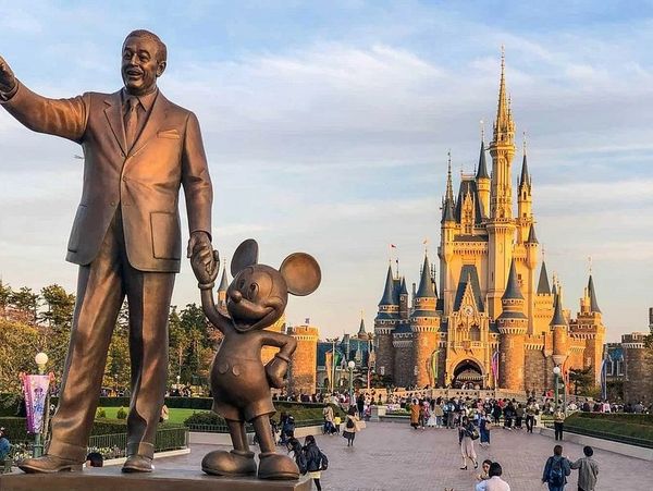 Statue of Walt Disney and Mickey Mouse in front of a grand castle with visitors.