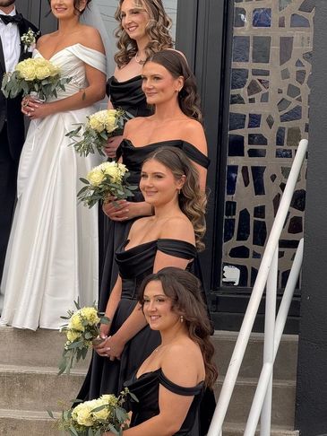 Bridesmaids in black dresses holding white flower bouquets on stairs.