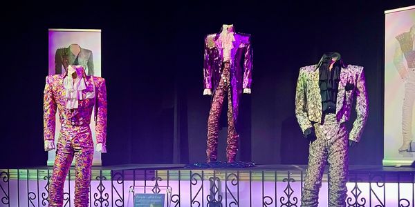 Three flamboyant, sequined suits displayed on mannequins in a dimly lit exhibition.