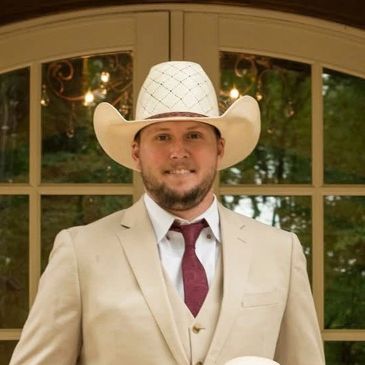 Man in beige suit and white cowboy hat standing indoors.