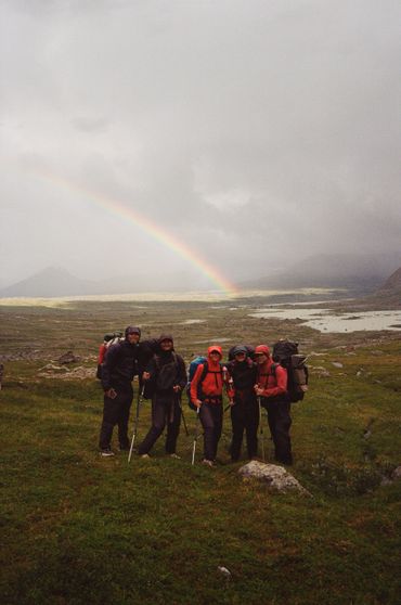 Five hikers stand together with a rainbow in the cloudy sky behind them.