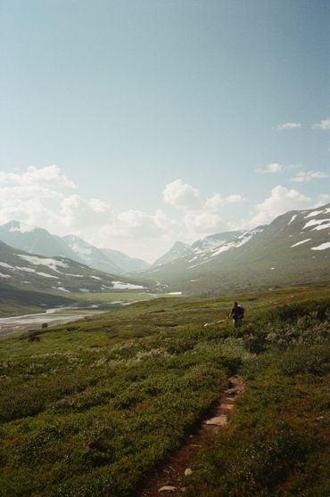 A lone hiker walks along a green mountain trail under a cloudy sky.