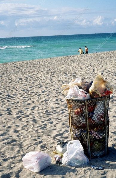 Overflowing trash bin on a sandy beach with two people near the water.