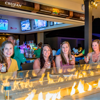 Four women enjoying drinks by a fire pit at a bar with TVs.