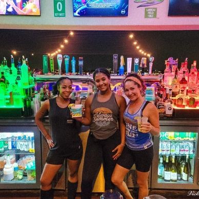 Three women posing happily behind a colorful bar with drinks in hand.