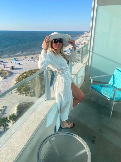 Woman in white dress and hat poses on balcony overlooking beach and ocean.