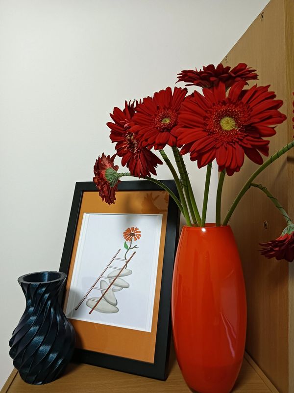 Red flowers in an orange vase with a framed flower artwork and a black textured vase.