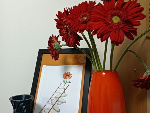 Red flowers in an orange vase with a framed flower artwork and a black textured vase.