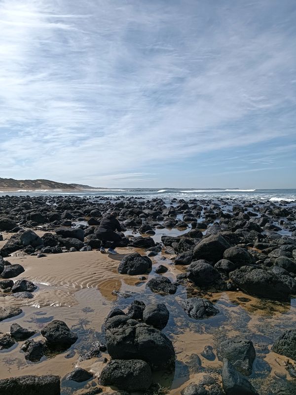 A person crouches among black rocks on a sandy beach under a cloudy sky.