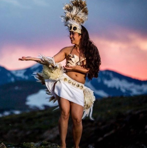 A woman in traditional Polynesian attire dancing outdoors at sunset.