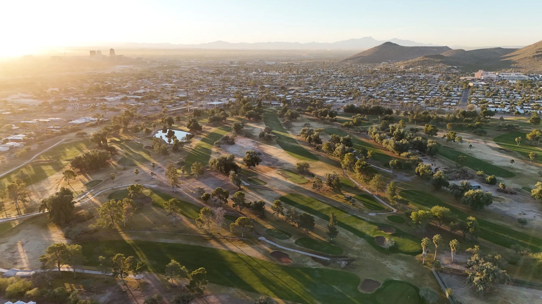 Aerial view of a golf course with a city and mountains in the background at sunset.