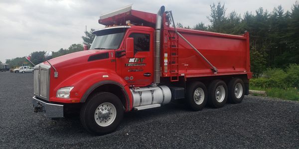 Red dump truck parked on gravel with trees in the background.