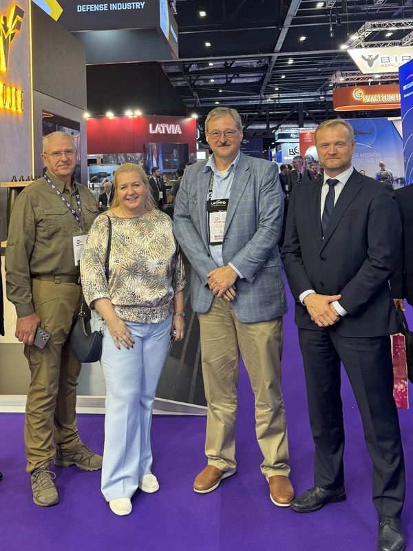 Four people posing at a defense industry event with Latvia booth visible.