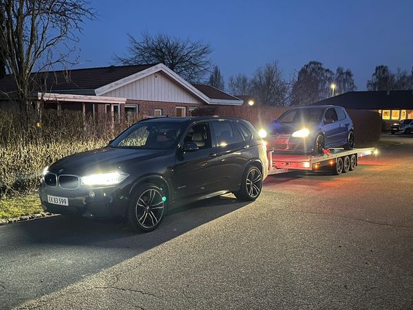 A black BMW SUV towing a small car on a trailer at dusk.