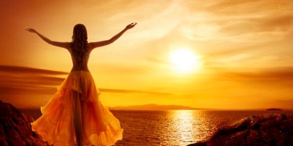 Woman in flowing dress embraces sunset by the sea.
