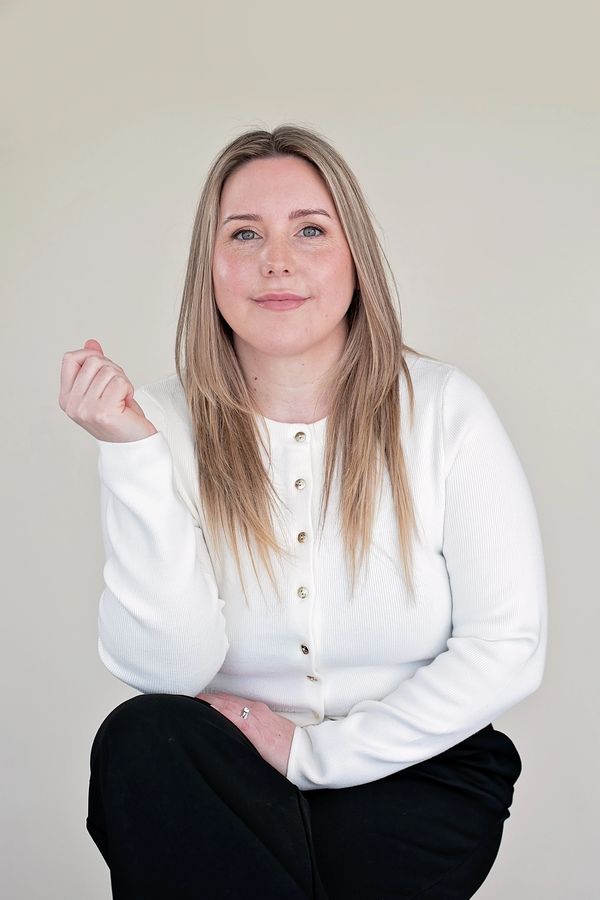 Confident woman in white cardigan and black pants poses against a plain background.
