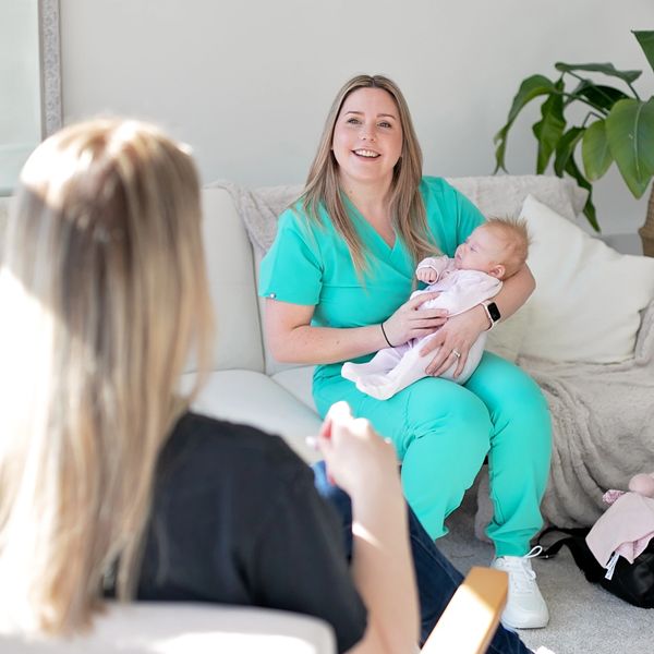 A nurse in scrubs happily holding a sleeping baby during a home visit.