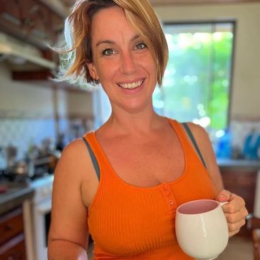 Smiling woman in an orange tank top holding a white mug in a kitchen.