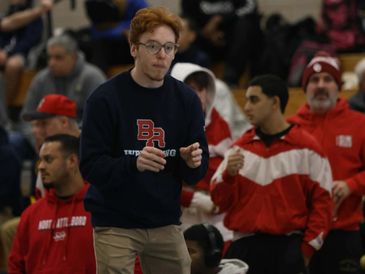 A young man in glasses and a sweatshirt stands focused among a crowd.