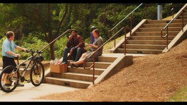 Three people talking by outdoor stairs, one with a bicycle.