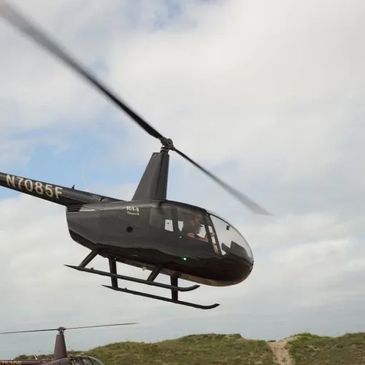 A black helicopter flying over grassy terrain under a cloudy sky.