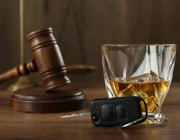 A glass of whiskey, car keys, and a judge's gavel on a wooden surface.