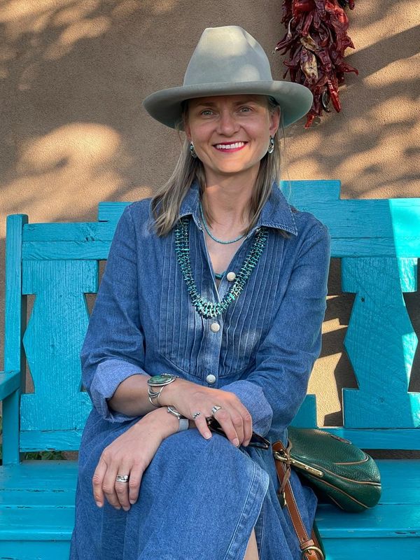 Woman in denim dress and hat sitting on a bright blue bench outdoors.