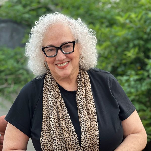 Smiling woman with white curly hair, black glasses, and leopard-print scarf outdoors.
